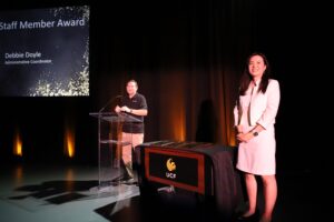 Nan Yu and Will Kinnally stand on stage at an awards event. A screen displays “Staff Member Award” and “Debbie Doyle, Administrative Coordinator.” A table with awards and a UCF logo is in the foreground.