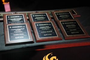 Several award plaques from the Nicholson School of Media, dated April 2025, are displayed on a table, recognizing achievements in staff, teaching, service, and research.