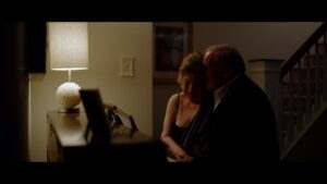 An older couple sits closely together in a dimly lit room near a piano, illuminated by a table lamp on a sideboard.