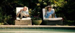 An older man and woman relax on poolside lounge chairs, holding drinks, surrounded by lush greenery and a large planter with flowers.