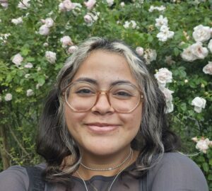 Karen Sanchez Crespo with glasses and wavy hair smiling in front of a bush with blooming light pink and white roses.