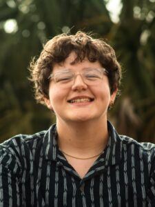 A person with short curly hair, glasses, and a striped shirt smiles outdoors with greenery in the background.