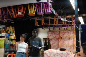 Two people stand and speak in a colorful, decorated room with "FRINGE" spelled out on the wall and upside-down chairs hanging from the ceiling.