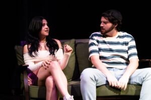 Two people sit on a green couch; the woman on the left looks upset and gestures with her hand, while the man on the right, wearing a striped shirt and cap, listens intently.