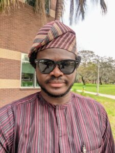 Tolulope Ogunrinde wearing sunglasses and a striped traditional outfit with a matching cap stands outside near a brick building and grassy area.