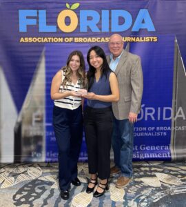 Three people stand together, with two holding an award plaque in front of a Florida Association of Broadcast Journalists backdrop.