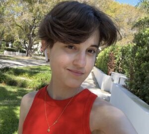 Victoria Deutsch with short brown hair wearing a red sleeveless top and a necklace takes a selfie outdoors in a sunny, tree-lined area.