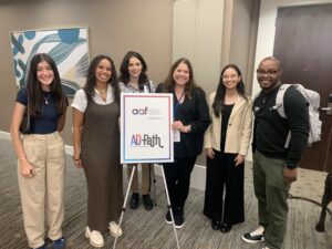 Six people stand indoors beside a sign that reads “AAF District 4 AD Path.” They are smiling and appear to be at a professional or educational event.