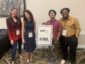 Four people stand indoors, smiling next to a sign that reads "AAF District 4 AD Path Student Conference." They are wearing name tags and casual to business-casual attire.