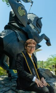 A person wearing graduation attire sits in front of a large black knight statue outdoors on a sunny day.
