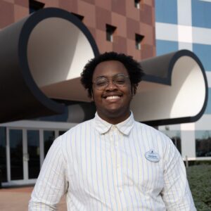 A person wearing glasses and a striped shirt stands smiling in front of a modern building with large, geometric black and white architectural features.