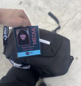 A person holds a WUSF Public Media badge labeled "MEDIA" with the name Kayla Kissel, News Intern, above a black bag on a light-colored surface.
