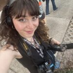 Woman with curly brown hair, wearing headphones and holding audio equipment, takes a selfie outdoors. She has a Nikon camera and press badges around her neck. Other people stand nearby.
