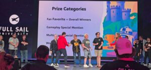 A group of people stand on stage at Full Sail University as prize categories are displayed on a large screen during an award event.