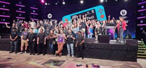 A large group of people pose and smile on stage at a gaming event, with colorful lights and screens displaying event logos in the background.