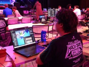 Person working on 3D modeling software at a laptop during a hackathon event; several other participants are visible in the background at tables with computers.