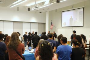 A group of people stand in a classroom facing speakers at the front, while one person appears on a video call projected on a screen above.