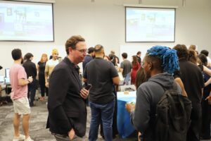 A group of people interact and network in a conference room with presentation screens and tables in the background.