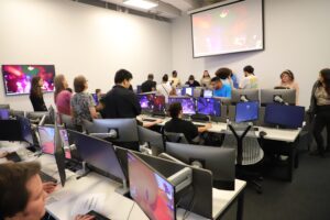 People work and interact at computer stations in a modern lab, with multiple monitors and a large screen displaying a video game on the wall.