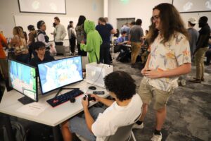 People gather in a room, some seated at a table with dual monitors playing a video game, while others stand and watch or talk in the background.