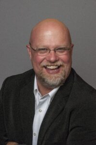 A bald man with glasses, a beard, and a mustache is smiling broadly. He is wearing a dark blazer over a white shirt, sitting in front of a plain gray background.