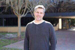 A man with short gray hair stands outdoors in front of a building, smiling at the camera. Leafless trees and a brick walkway are visible in the background.
