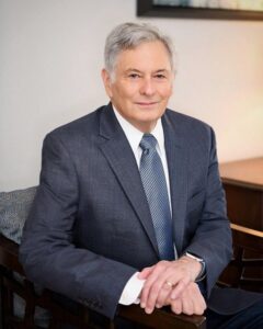 An older man with gray hair wearing a navy suit, white shirt, and patterned tie sits on a wooden chair, hands folded, looking at the camera.