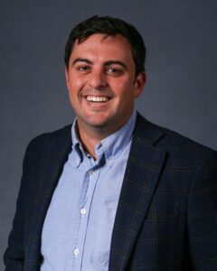 A man wearing a blue button-up shirt and a dark plaid blazer stands smiling in front of a plain dark background.