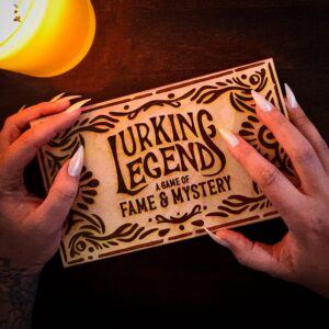 Two hands with long, metallic nails hold a boxed game titled "Lurking Legend: A Game of Fame & Mystery" on a dark surface next to a lit candle.