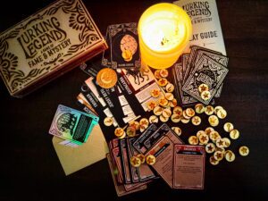 A tabletop game called "Lurking Legend" is spread out, with cards, tokens, a play guide, an envelope, a candle, and the game box visible on a dark surface.