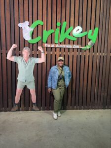 A person stands next to a life-sized cutout of Steve Irwin under a sign that reads "Crikey I'm at Australia Zoo," with a wooden wall in the background.