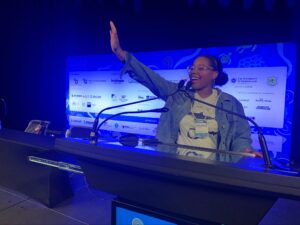A woman stands at a podium on a stage, smiling and raising one hand, with sponsor logos displayed on a blue backdrop behind her.