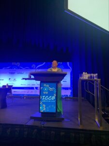 A woman stands at a podium on a stage, presenting at ICCB 2023. The background features event sponsors' logos and a table with water bottles is visible to the side.
