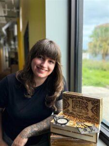 A person with brown hair and a black shirt smiles next to stacked board game boxes labeled "Lurking Legend" by a window with a green outdoor view.