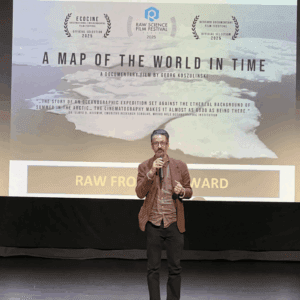 A man stands on stage with a microphone in front of a screen displaying information about the documentary film "A Map of the World in Time" and its festival awards.