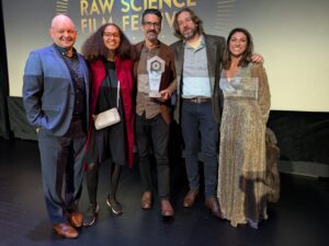 Five people stand together on stage at the Raw Science Film Festival, with one person in the center holding a trophy.