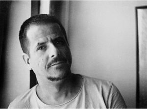 A man with short hair and a goatee is looking at the camera while standing near a window. The image is in black and white.
