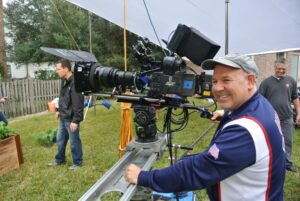 A camera operator adjusts a large film camera on a track outdoors while several people stand in the background on a grassy lawn.
