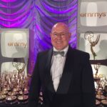 A man in a tuxedo stands in front of a display of Emmy Awards and television props with purple lighting in the background.