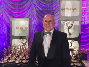 A man in a tuxedo stands in front of a display of Emmy Awards and television props with purple lighting in the background.
