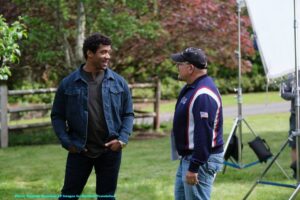 Two men stand on grass outdoors, smiling and talking. There is a wooden fence, trees, and professional lighting equipment in the background.