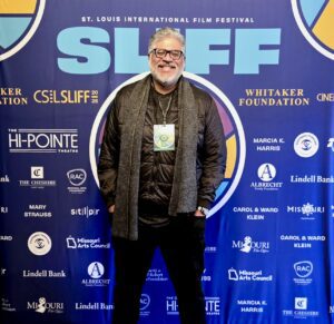 A man stands smiling in front of a blue St. Louis International Film Festival backdrop, wearing glasses, a scarf, and a black jacket with a festival badge around his neck.