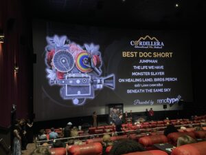 A movie theater screen displays nominees for Best Doc Short at the Cordillera International Film Festival while people stand near the stage in front of seated audience members.