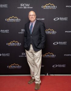 A man in a blazer, dress shirt, and khaki pants stands on a red carpet in front of a Cordillera International Film Festival backdrop.