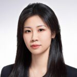 A woman with straight black hair wearing a black top and a necklace, facing forward against a plain light background.