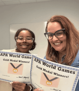 Two women smiling and holding certificates that say "APA World Games Gold Medal Winners!" in a room with white walls.