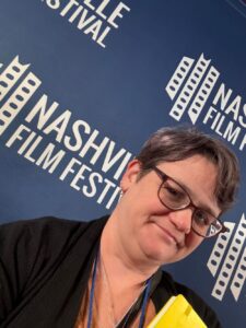 A person wearing glasses poses in front of a blue Nashville Film Festival backdrop, holding a yellow item.