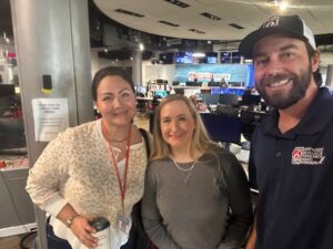 Three people stand and smile for a photo inside a busy newsroom with desks, monitors, and a "Weather Center" sign visible in the background.