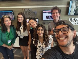 Six people pose and smile for a group selfie in an office setting, standing in front of wall-mounted screens and a “True Crime Network” sign.