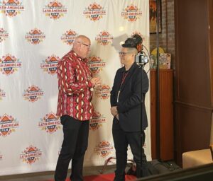 Two men stand and talk on a red carpet in front of a step-and-repeat banner for the Oklahoma Latino American Film Festival. A camera and light are set up in front of them.
